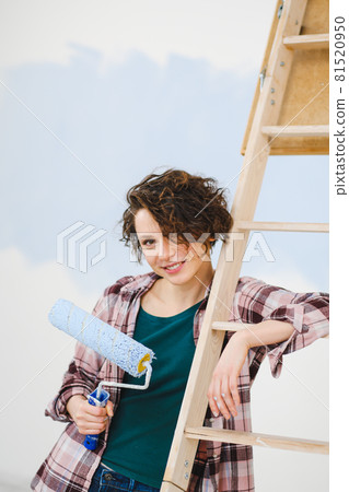 A young beautiful woman painter designer paints a white wall with blue paint with roller. Plaid shirt, short hair and jeans. 81520950