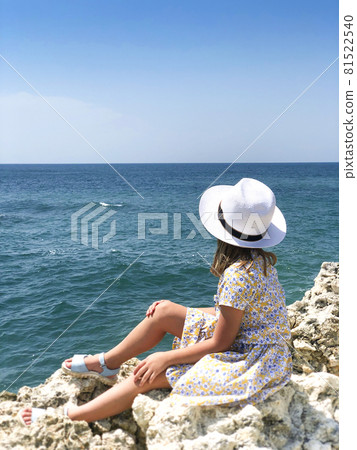 Little girl sitting on rocky shore while looking at sea during summer vacation with family on seaside 81522540