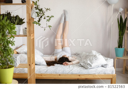 Young slim brunette woman in casual clothes lying down on wooden bed with legs raised up on wall at home Young slim brunette woman in casual clothes lying down on wooden bed with legs raised up on wall at home 81522563