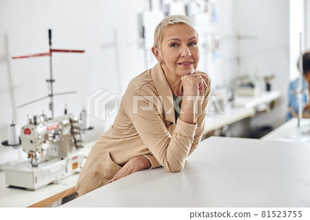 Elegant mature woman in beige jacket leans onto white cutting table in sewing workshop 81523755
