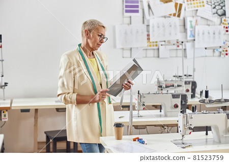 Mature lady looks at open folder standing nera workplace with sewing machine Mature lady looks at open folder standing nera workplace with sewing machine 81523759