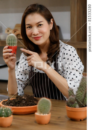 Beautiful Asian woman wearing apron showing a small pod of cactus with happy and proud for her hobby and home gardening in free time 81533280