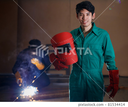 smart confident young Asian man wearing a factory uniform holding a welding helmet posing smiling to a camera with soft background on his colleague working on welding smart confident young Asian man wearing a factory uniform holding a welding helmet posing smiling to a camera with soft background on his colleague working on welding 81534414