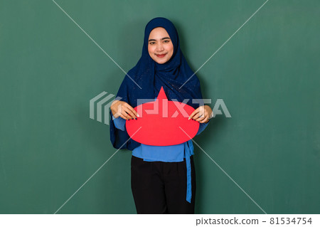confident and happy woman concept. Pretty attractive Muslim lady dressed beautifully wearing blue hijab standing and holding a red blank speech bubble over green background confident and happy woman concept. Pretty attractive Muslim lady dressed beautifully wearing blue hijab standing and holding a red blank speech bubble over green background 81534754