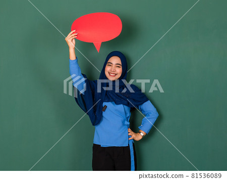 confident and happy woman concept. Pretty attractive Muslim lady dressed beautifully wearing blue hijab standing and holding a red blank speech bubble over green background confident and happy woman concept. Pretty attractive Muslim lady dressed beautifully wearing blue hijab standing and holding a red blank speech bubble over green background 81536089