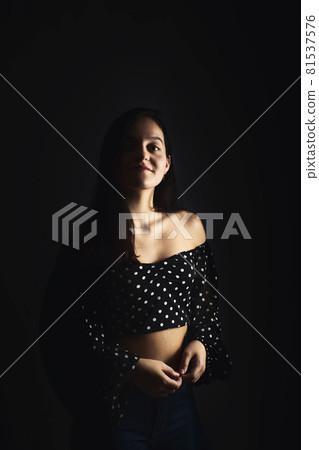 Half-length portrait of a young girl with a smile looking towards the camera, on a dark studio background Half-length portrait of a young girl with a smile looking towards the camera, on a dark studio background 81537576