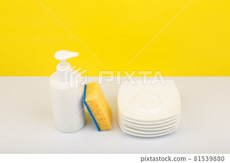 Dishwashing detergent in white bottle, pile of saucers and cleaning sponge against yellow background 81539880