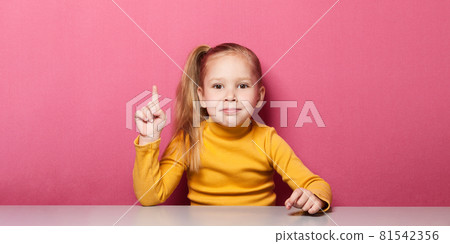 Girl is sitting at the table and shows her index fingers up Girl is sitting at the table and shows her index fingers up 81542356
