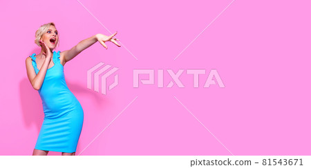 Portrait of beautiful blonde woman in blue dress posing on pink studio background, pointing finger to empty space 81543671