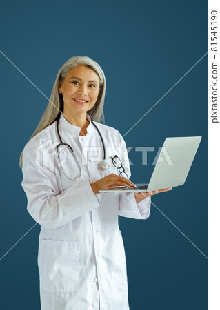 Cheerful mature lady doctor in white robe uses modern laptop standing on blue background Cheerful mature lady doctor in white robe uses modern laptop standing on blue background 81545190