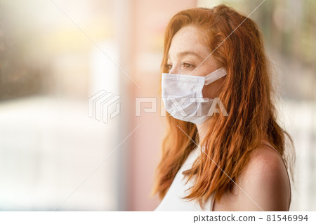 Young woman is standing and looking from window hall with a protection mask on her face. Corona virus outbreaking. Young woman is standing and looking from window hall with a protection mask on her face. Corona virus outbreaking. 81546994