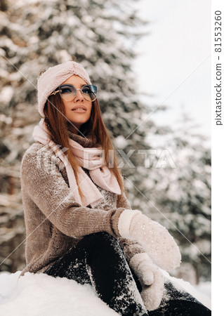 A girl in a sweater and glasses in winter sits on a snow-covered background in the forest 81553260