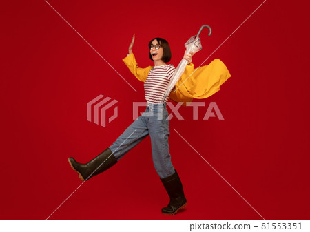 Autumn weather. Young woman wearing raincoat and gumboots struggling wind and walking over red background 81553351