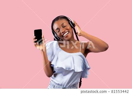 Cool black lady in headphones listening to music, showing smartphone with empty screen on pink background, mockup Cool black lady in headphones listening to music, showing smartphone with empty screen on pink background, mockup 81553442