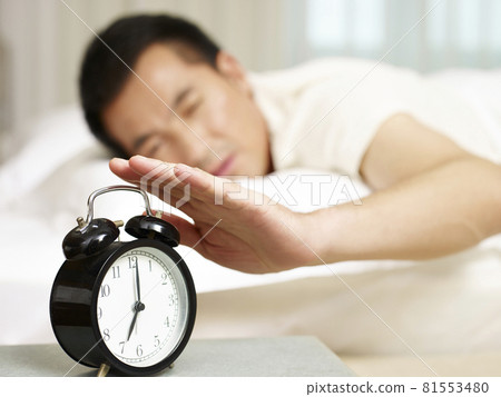 asian man lying in bed trying to stop ringing alarm clock, focus on the clock asian man lying in bed trying to stop ringing alarm clock, focus on the clock 81553480
