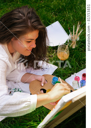 Artist painting on the easel outdoors in the garden. Open air outdoor art workshop. Draw on the canvas with brush and palette sitting on the grass during a picnic in a park 81553831