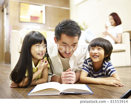 asian father and two children lying on front on floor reading book mother relaxing in background. asian father and two children lying on front on floor reading book mother relaxing in background. 81553914