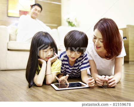asian mother and two children lying on floor playing with ipad father watching in background 81559678