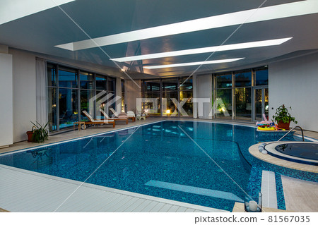 Luxurious indoor pool with jacuzzi bathtub and loungers against backdrop of green evening park outside windows 81567035