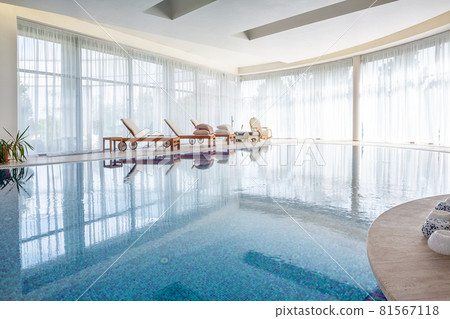 Swimming pool with wooden loungers and wicker rocking chair in spacious room of luxurious mansion 81567118