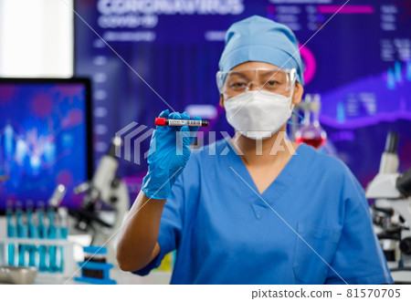 Female medical people wearing a hygiene face mask and protective glass staying in laboratory room, holding sample test tube with lab equipment in background and looking to camera Female medical people wearing a hygiene face mask and protective glass staying in laboratory room, holding sample test tube with lab equipment in background and looking to camera 81570705