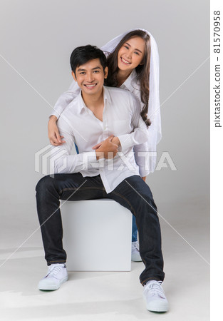 Young attractive Asian couple wearing white shirt and veil. woman holding man from the back and smiling against white background. Concept for pre wedding photography. Isolated Young attractive Asian couple wearing white shirt and veil. woman holding man from the back and smiling against white background. Concept for pre wedding photography. Isolated 81570958