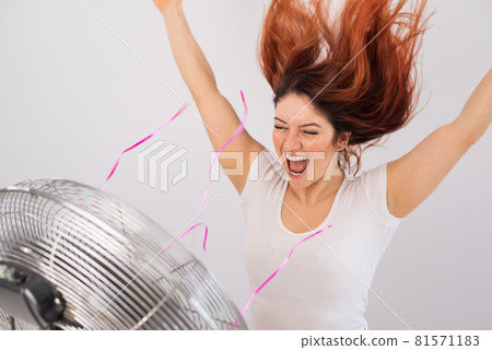 Joyful caucasian woman enjoying the wind blowing from an electric fan on a white background. 81571183