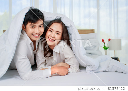 Young happy attractive Asian couple lying in bed together looking at camera with blanket covering them. White curtain background. Concept for love and happy relationship 81572422