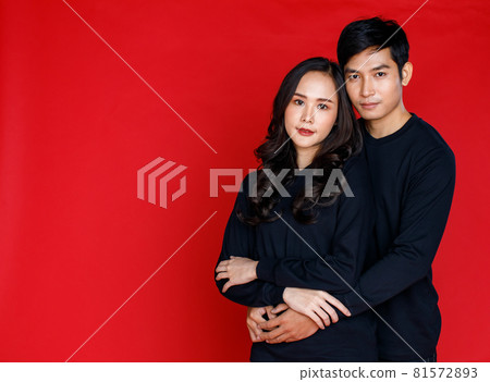 Young attractive Asian couple wearing black shirt and jeans holding each other against red background. Concept for pre wedding photography. Isolated Young attractive Asian couple wearing black shirt and jeans holding each other against red background. Concept for pre wedding photography. Isolated 81572893