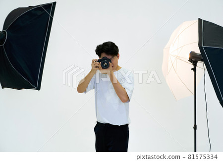 Young asian photographer looks through the viewfinder while shooting with the camera. Working atmosphere in the photo studio. Young asian photographer looks through the viewfinder while shooting with the camera. Working atmosphere in the photo studio. 81575334