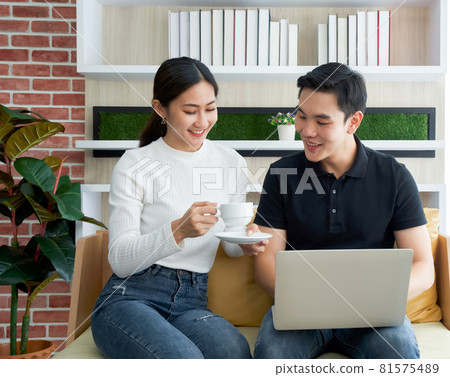 Asian woman in white long sleeve t-shirt handed a cup of coffee to her boyfriend while the man held laptop computer. Young lovers spend time together on holidays in the living room. 81575489