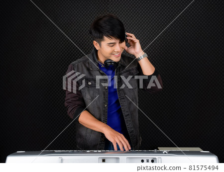 Young asian man with headphones playing an electric keyboard in front of black soundproofing walls. Musicians producing music in professional recording studio. 81575494