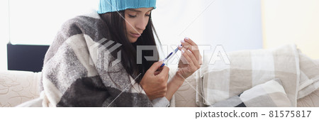 Woman in hat sitting under covers and looking at thermometer 81575817