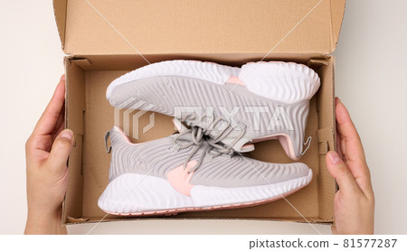 female hands are holding a brown cardboard box with gray sports sneakers on a white table, inspecting an online order 81577287
