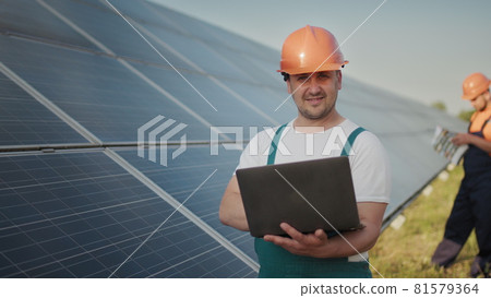 Engineer standing with Solar cell panels looking monitor of the Electric power value made from the solar cell. Man holding laptop in her hands standing on field with solar panels. Solar energy Engineer standing with Solar cell panels looking monitor of the Electric power value made from the solar cell. Man holding laptop in her hands standing on field with solar panels. Solar energy 81579364