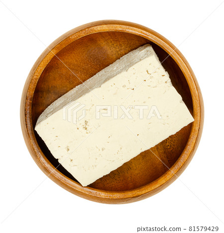 Block of white tofu, in a wooden bowl. Bean curd, coagulated soy milk, pressed into white blocks of different softness. Component of Asian cuisine and meat substitute. Close-up from above, over white. 81579429