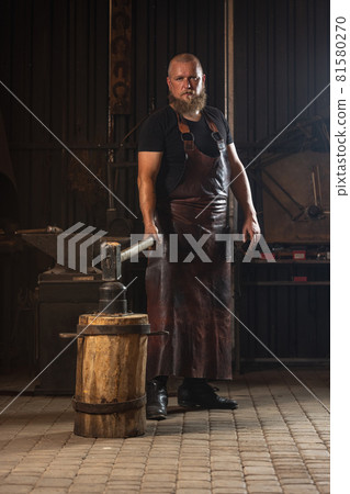 Full ength portrait of bearded man, blacksmith working on creating handmade metal product at family smithy. Concept of labor, retro professions, power, beauty Full ength portrait of bearded man, blacksmith working on creating handmade metal product at family smithy. Concept of labor, retro professions, power, beauty 81580270