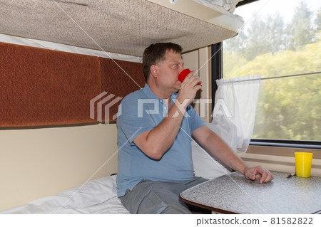 A man in a compartment car drinks tea with pleasure from a red mug and looks out the window 81582822