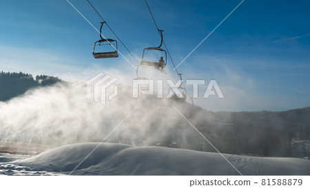 Stunning views of ski lift, artificial snow powder in morning sun at ski resort. Skiers move on chair lift, backdrop of blue sky, winter forest, white snow. Perfect weather for skiing, snowboarding 81588879