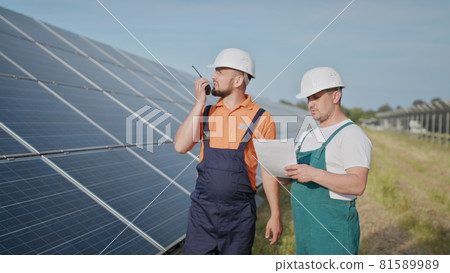 Coworking engineers on solar plant. Adult men in hardhats, An employee of a power plant transmits commands by walkie-talkie. Transportation of clean energy. Clean energy. Sunny batteries. Coworking engineers on solar plant. Adult men in hardhats, An employee of a power plant transmits commands by walkie-talkie. Transportation of clean energy. Clean energy. Sunny batteries. 81589989