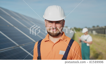 Happy caucasian adult engineer in helmet smiling at camera at solar power station outside. Ecological industry. Solar farm concept. An employee of a power plant transmits commands by walkie-talkie 81589996