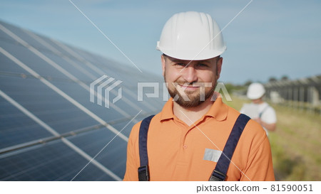 Happy caucasian adult engineer in helmet smiling at camera at solar power station outside. Ecological industry. Solar farm concept. An employee of a power plant transmits commands by walkie-talkie Happy caucasian adult engineer in helmet smiling at camera at solar power station outside. Ecological industry. Solar farm concept. An employee of a power plant transmits commands by walkie-talkie 81590051