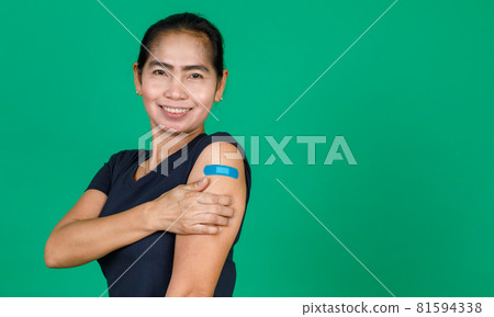 Middle aged Asian woman smiling showing her arm with bandage patch showing she got vaccinated for Covid 19 virus on green background. Concept for Covid 19 vaccination 81594338