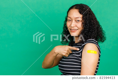 Asian middle aged woman smiling and pointing at her arm with bandage patch showing she got vaccinated for Covid 19 virus on green background. Concept for Covid 19 vaccination 81594413