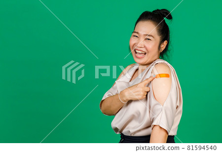 Asian middle aged woman smiling and pointing at her arm with bandage patch showing she got vaccinated for Covid 19 virus on green background. Concept for Covid 19 vaccination Asian middle aged woman smiling and pointing at her arm with bandage patch showing she got vaccinated for Covid 19 virus on green background. Concept for Covid 19 vaccination 81594540
