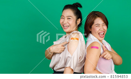 Two middle aged Asian women showing their arms with bandage patch showing they got vaccinated for Covid 19 virus on green background. Concept for Covid 19 vaccination 81594853