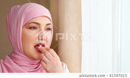 Young woman in hijab drinks white capsule with water while standing by the window 81597823