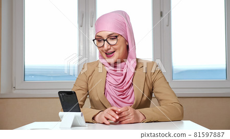 woman in a hijab and glasses sits at a table and talks by video call on the phone woman in a hijab and glasses sits at a table and talks by video call on the phone 81597887
