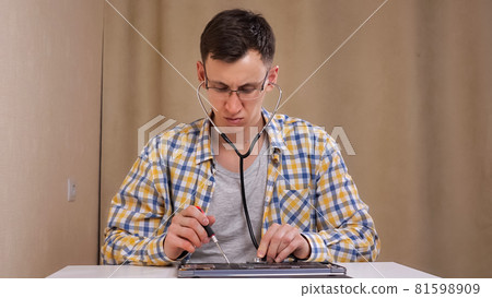 man with glasses examines a disassembled laptop and listens with a phonendoscope 81598909