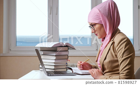 Muslim woman writes with a pen in a notebook looking into a laptop Muslim woman writes with a pen in a notebook looking into a laptop 81598927
